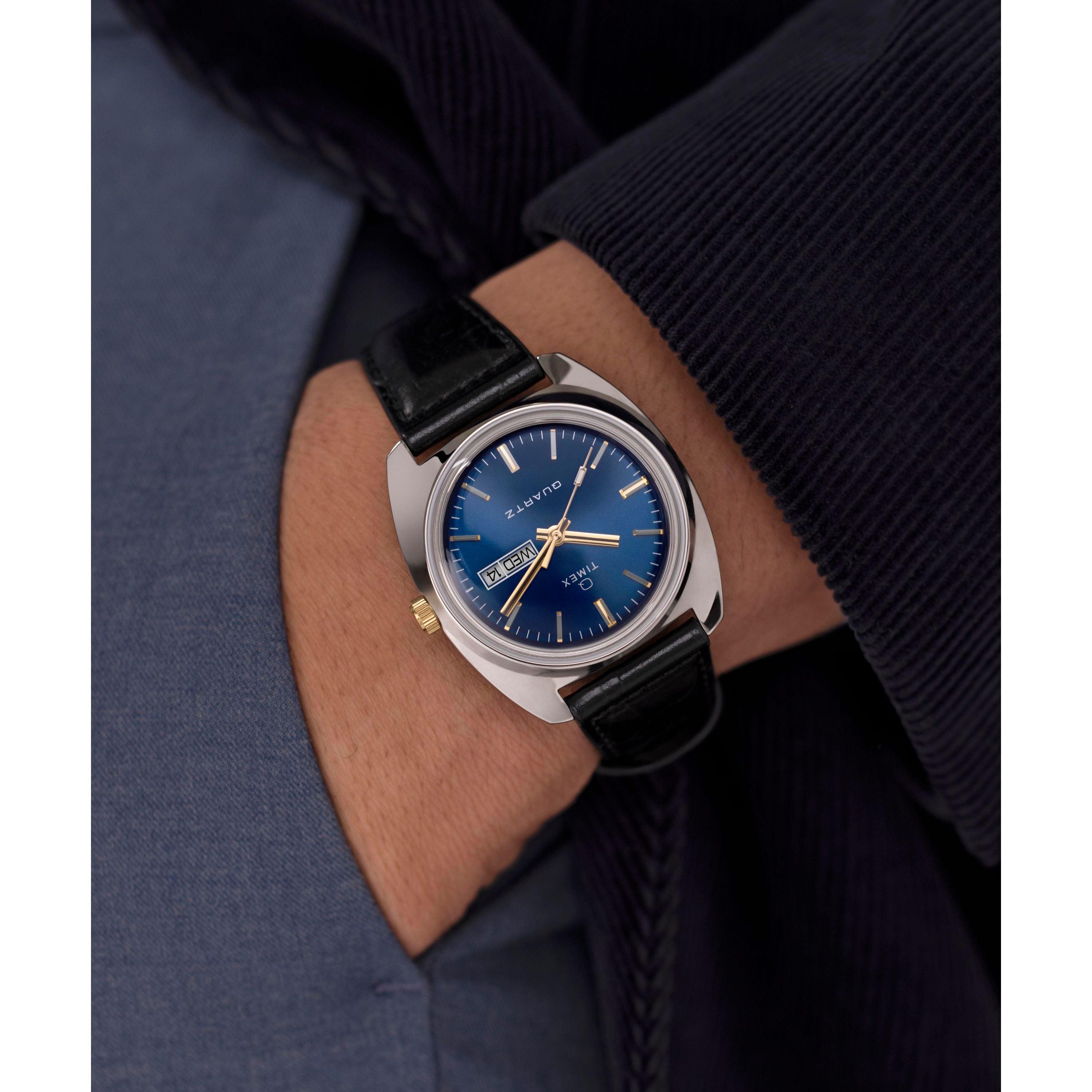 A person in a dark jacket and blue shirt wears the Timex G SS QZ SW 1978 DAY DTE 37.5 BLU DL 25, a retro dress watch with blue sunray dial, silver case, gold crown, black leather strap, and day-date display.