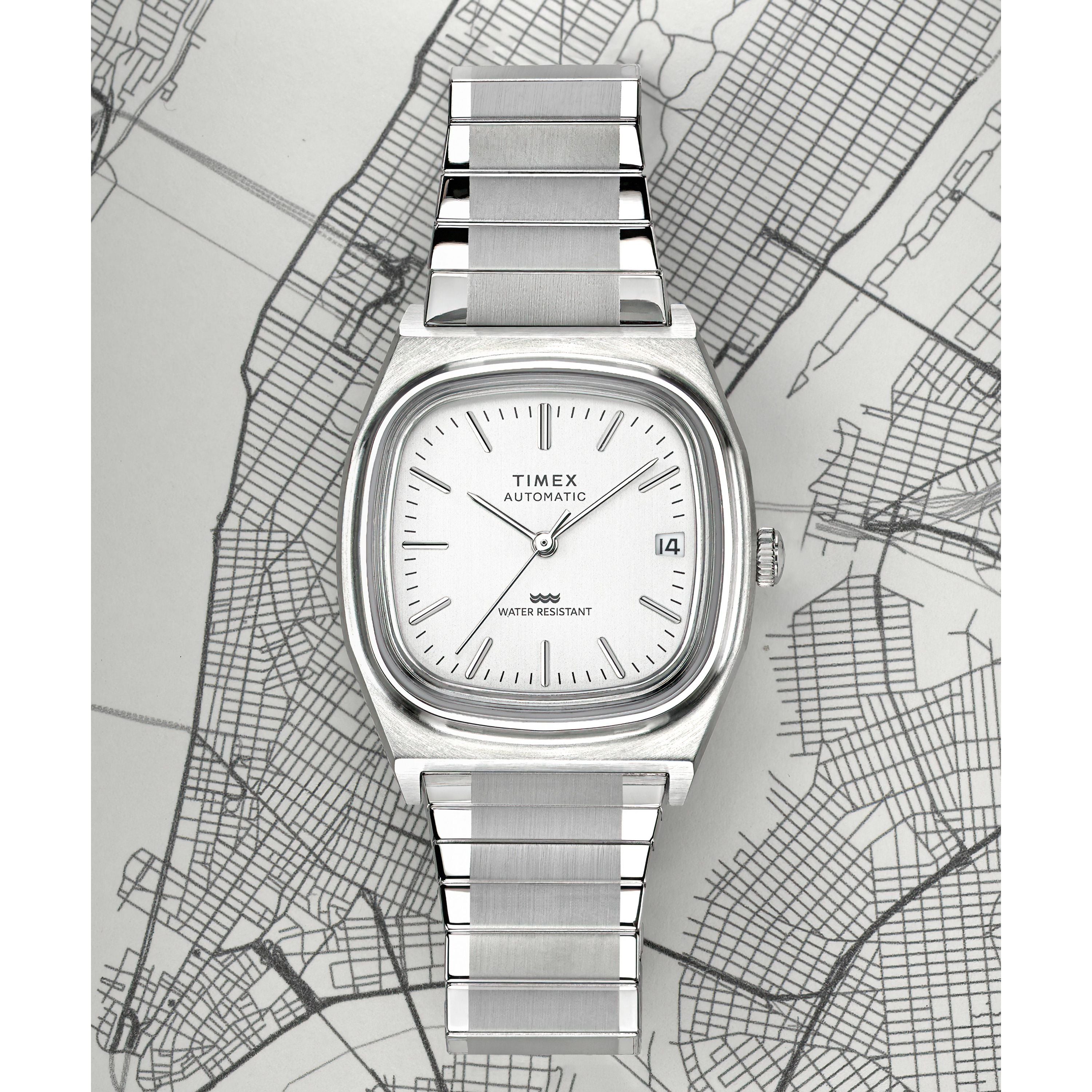 The Timex G SS AU BW E LINE REISSUE 34 WHITE DL 25 wristwatch, with a silver metal band and white dial, is shown on a grayscale city map. It features simple markers, a date at 3 o’clock, and Japanese automatic movement.