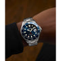 A person wearing a Timex G SS QZ BW DEEP WTR MERDIAN 44 BLU DL 25 dive watch, made from recycled stainless-steel, with a blue face, white markers, metal band, 200m water resistance, rotating bezel, and luminous details.