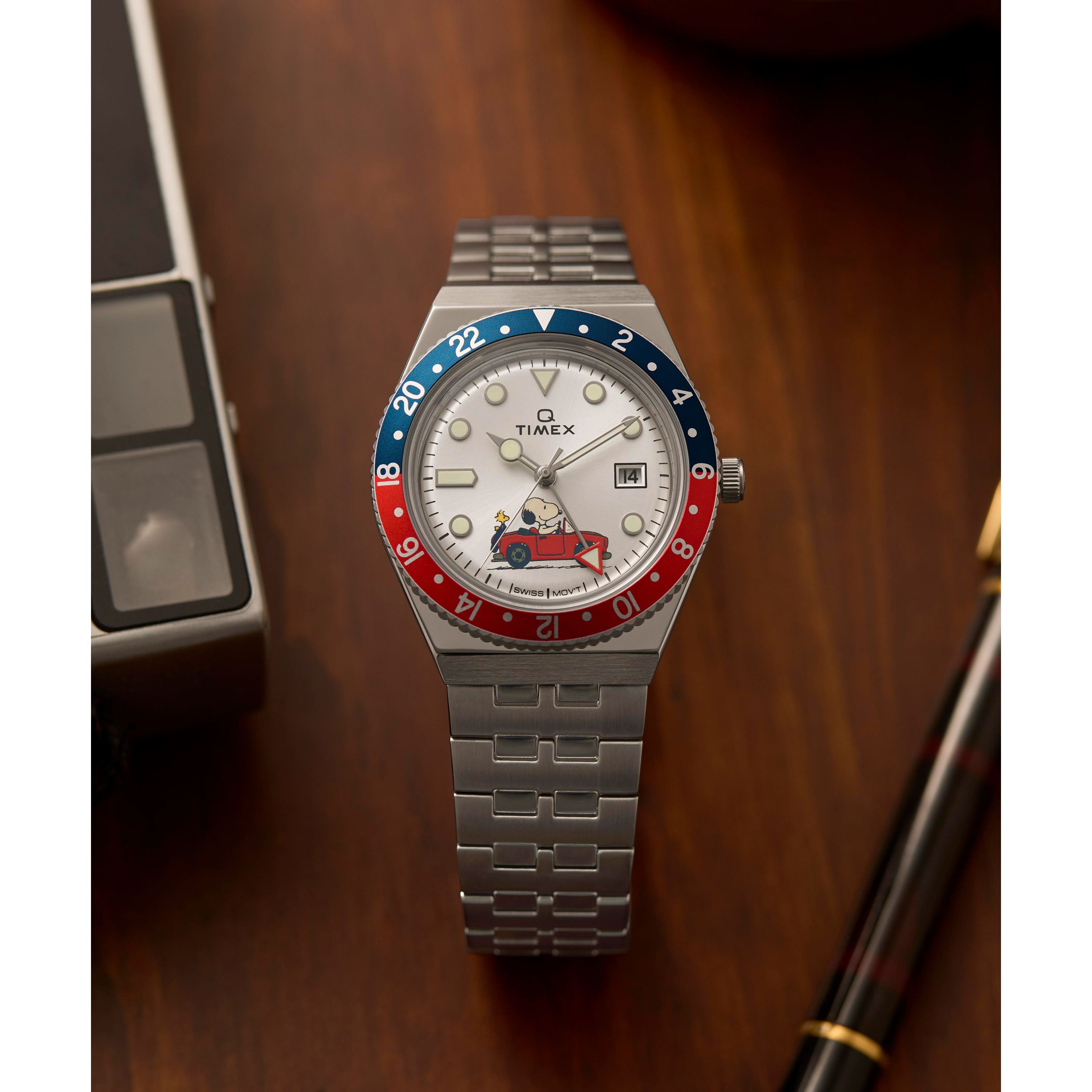 The Timex G SS QZ PEANUTS GMT 38 SLVR DL SS BLT 25, featuring a stainless-steel case, silver metal band, white dial, and a blue-red bezel, rests on a wooden surface beside a fountain pen and a vintage electronic device.