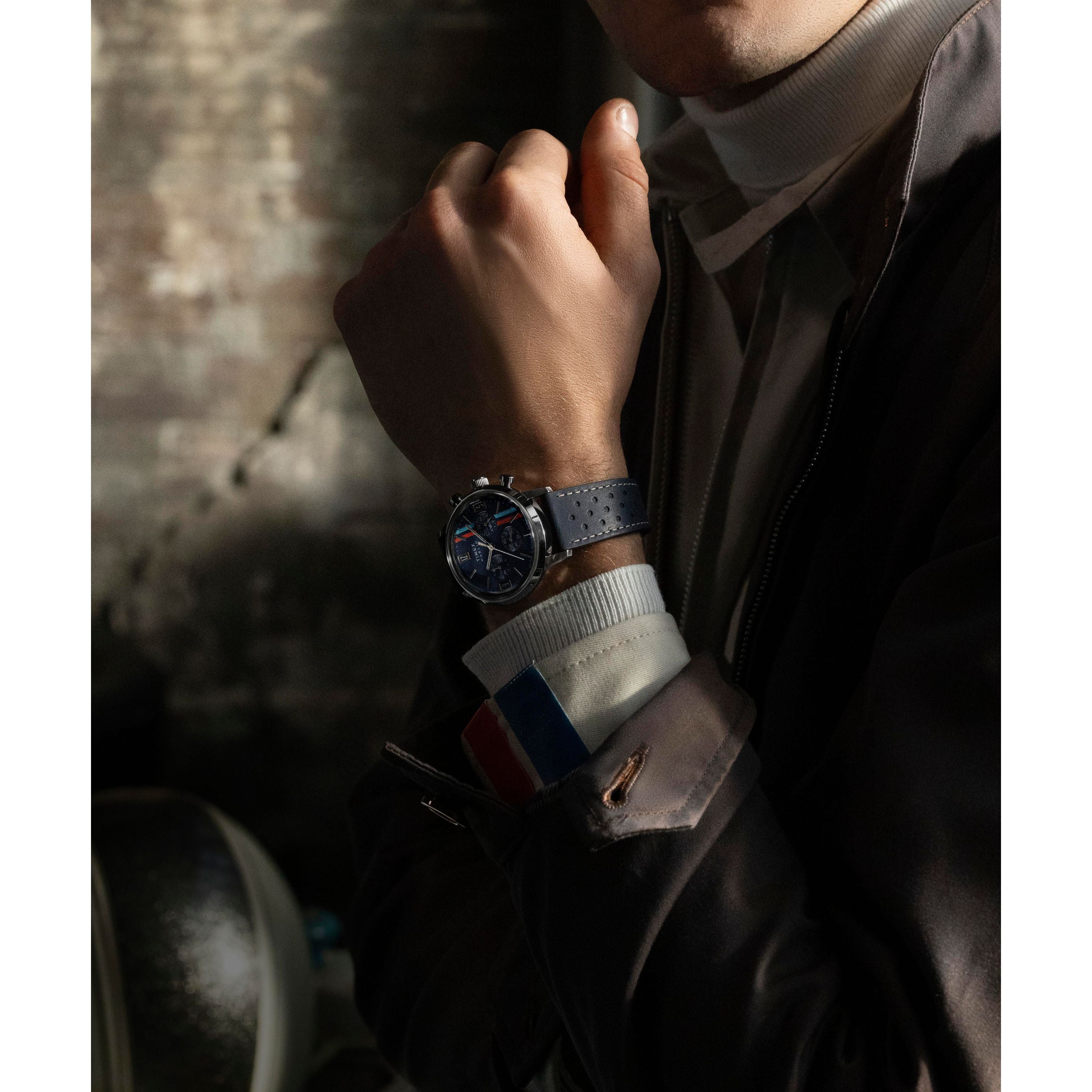 A person in a brown jacket and white turtleneck showcases the Timex Marlin® Chronograph Tachymeter with a perforated strap. Lighting highlights the watch and the French flag cuff detail against a dark, textured background.