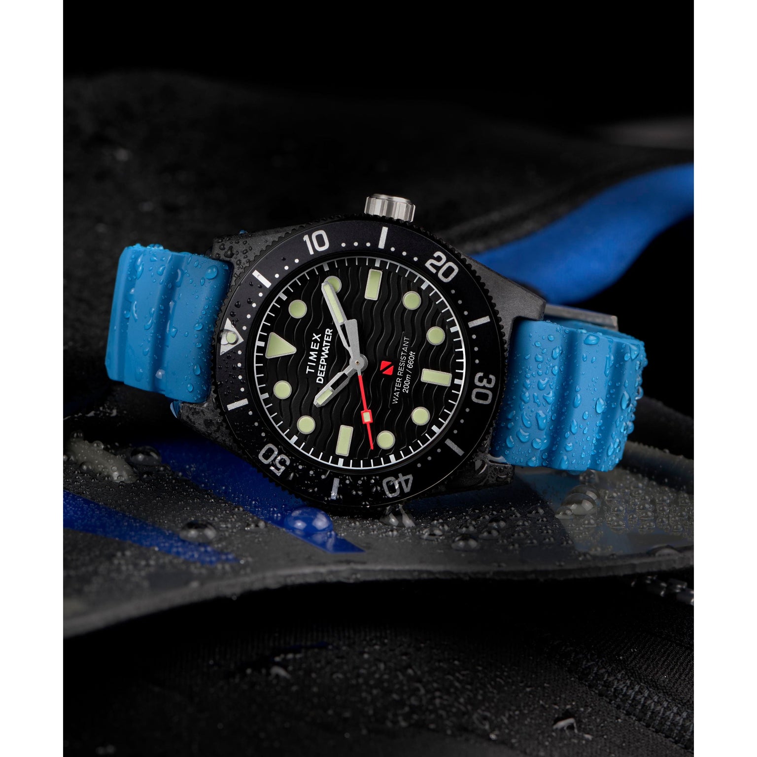 A Timex Deepwater Reef 200 XCF dive watch with a blue rubber strap and black bezel, covered in water droplets, lies on a wet black surface with blue accents. Its dial features bold white markers, hands, and 200m water resistance.