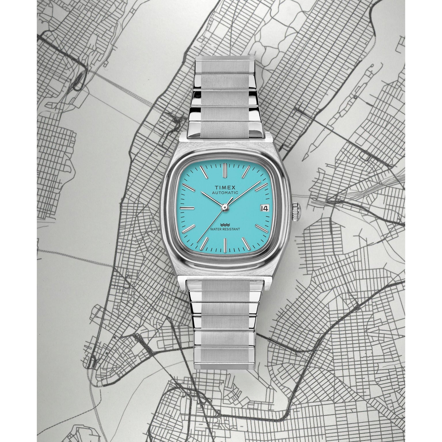 A Timex Automatic 1983 E Line dress watch with a turquoise face and metal band is displayed on a black-and-white New York City map, showing the date and time with the cityscape visible beneath it.