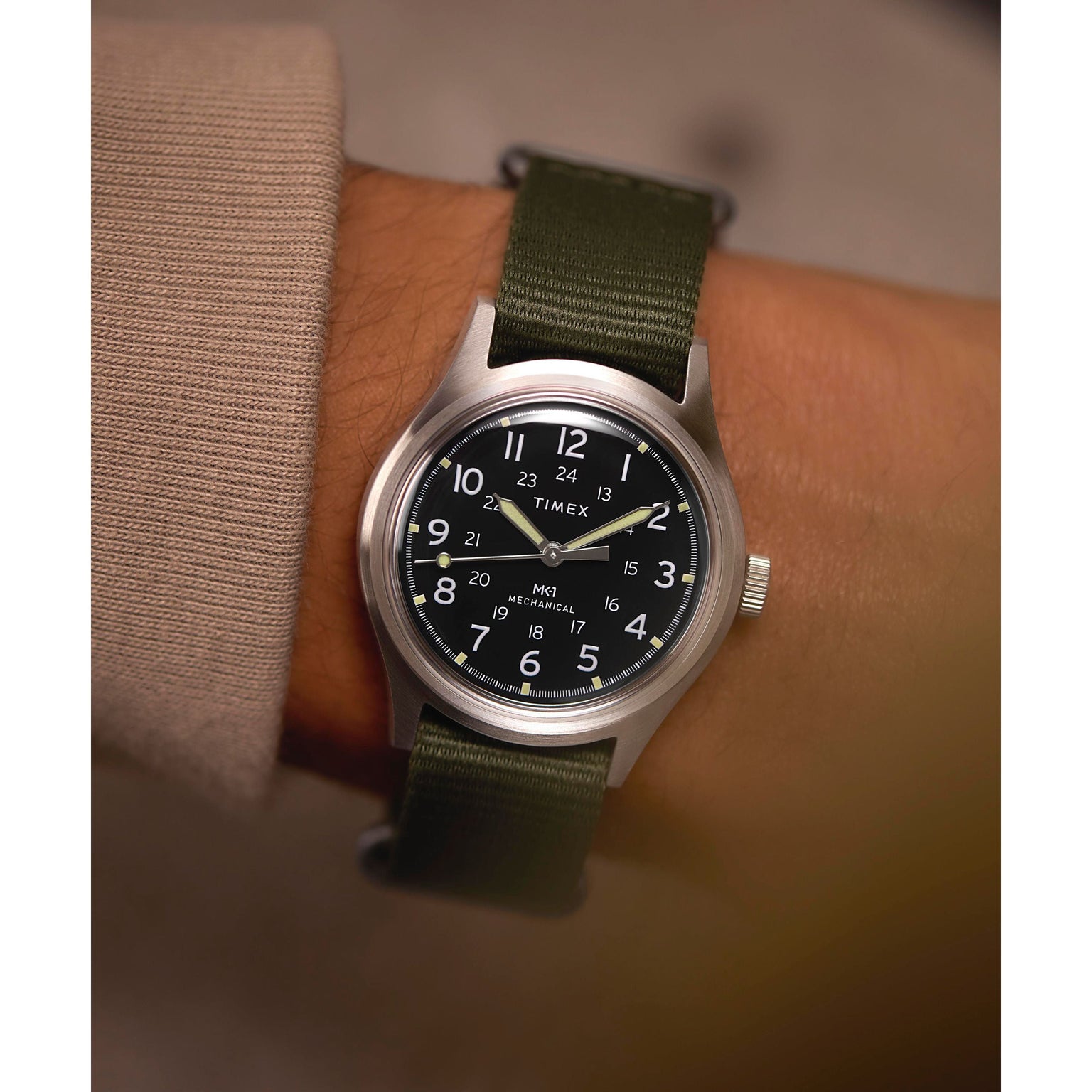 A person wears a Timex G SS MW SW MK1 36 BLK DL GRN STP 25 with a black dial, white numerals, and an olive green recycled strap, showing 10:09 over a beige long-sleeve shirt.