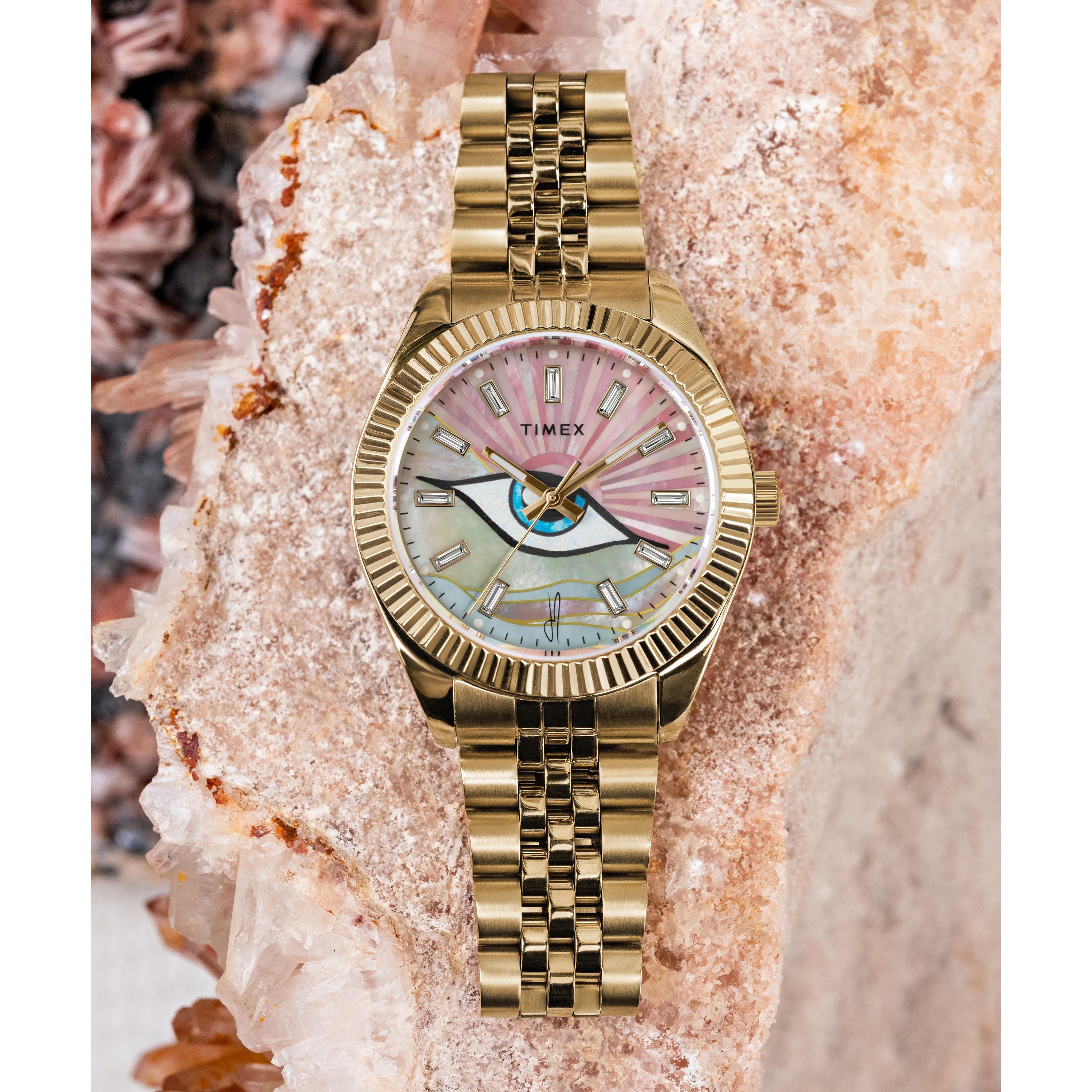 A Timex x Jacquie Aiche wristwatch with a gold link band and multicolored mother-of-pearl dial sits on a large crystal formation in pink and beige tones.