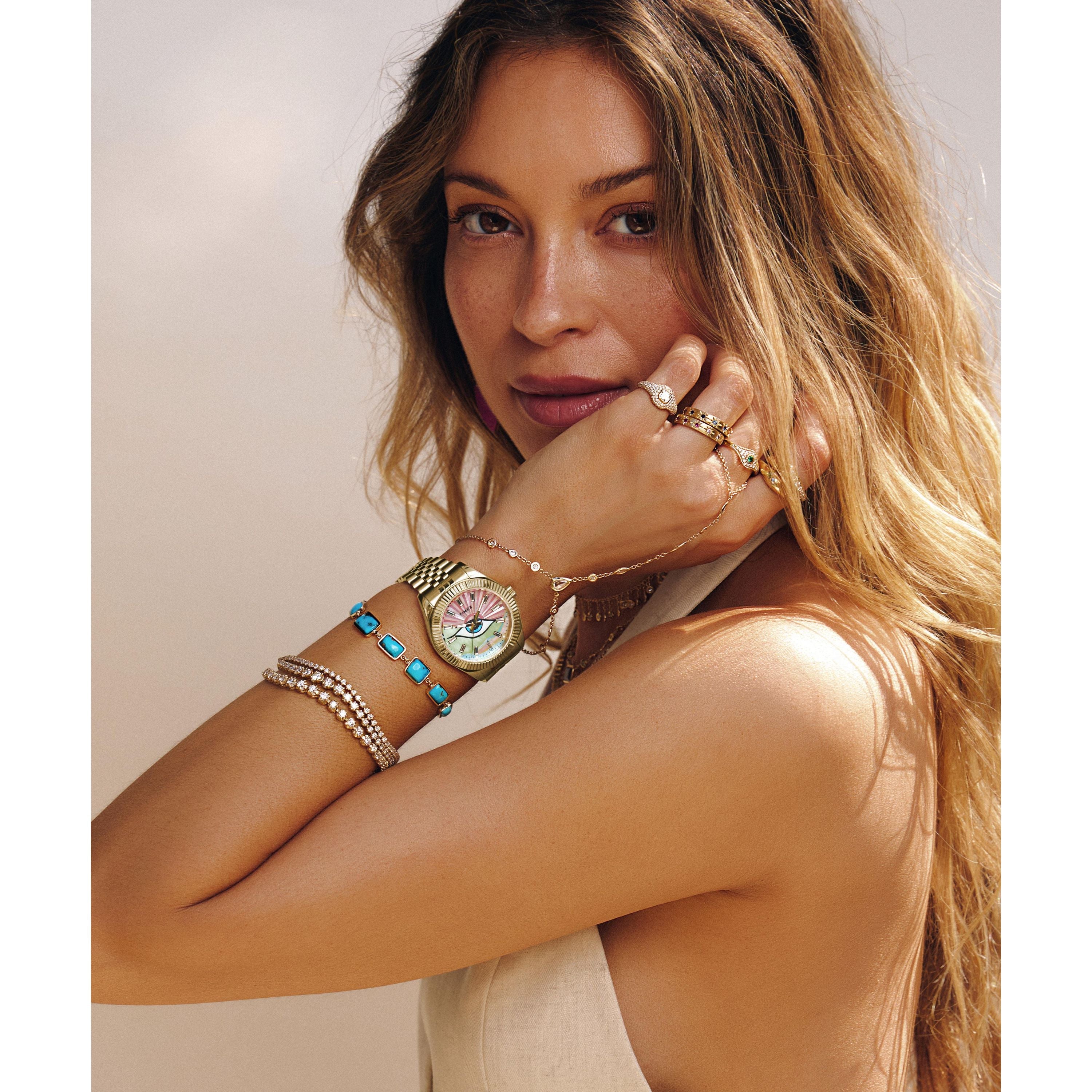 A woman with long, wavy blonde hair poses with her arm raised, showcasing layered bracelets, rings, and a Timex x Jacquie Aiche watch by Timex. She wears a sleeveless beige top and looks at the camera against a soft, neutral background.