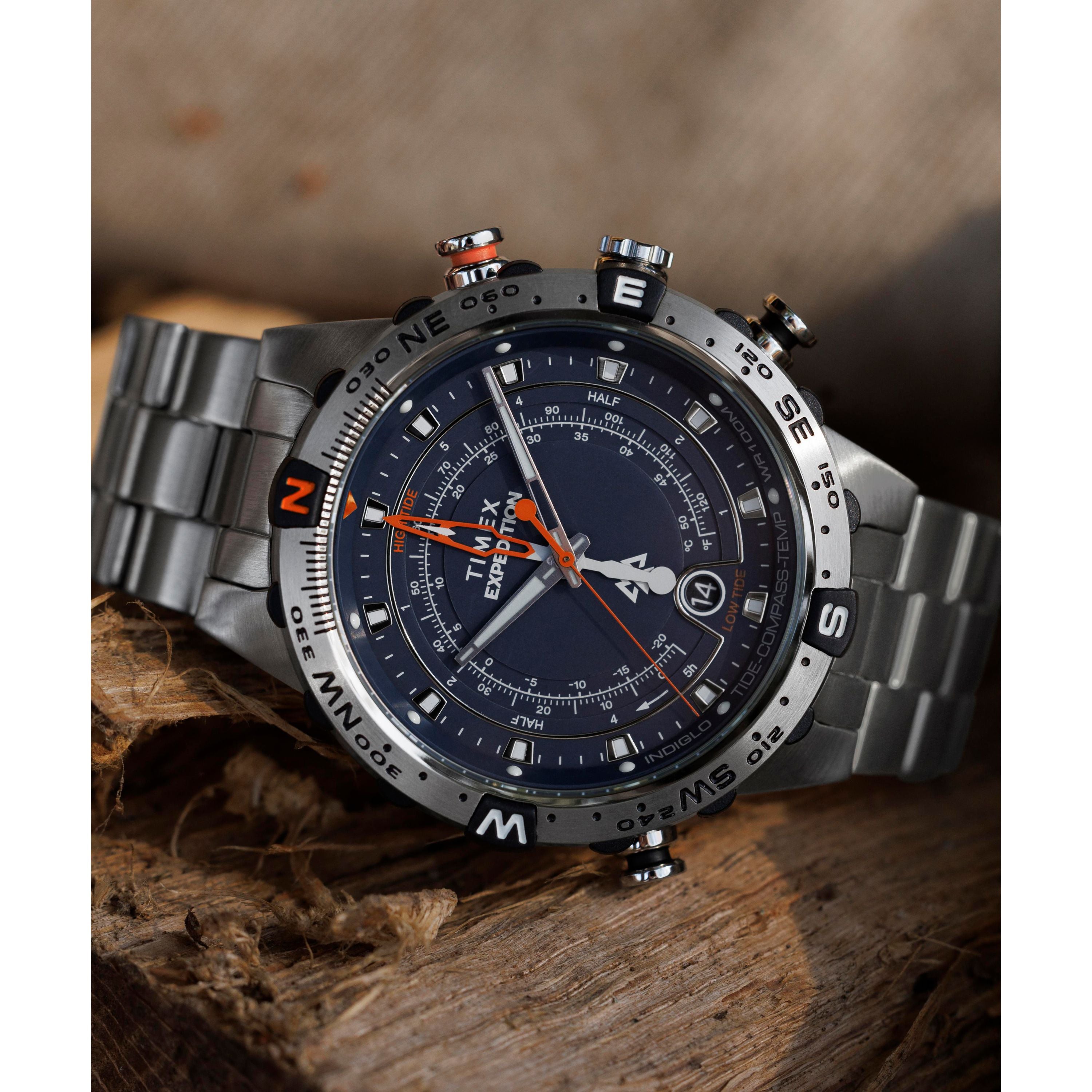 The Timex Expedition Tide-Temp-Compass watch, with its black dial, compass bezel, and orange accents, rests on rough wooden planks. Multiple dials and date display add to its rugged yet refined appeal.