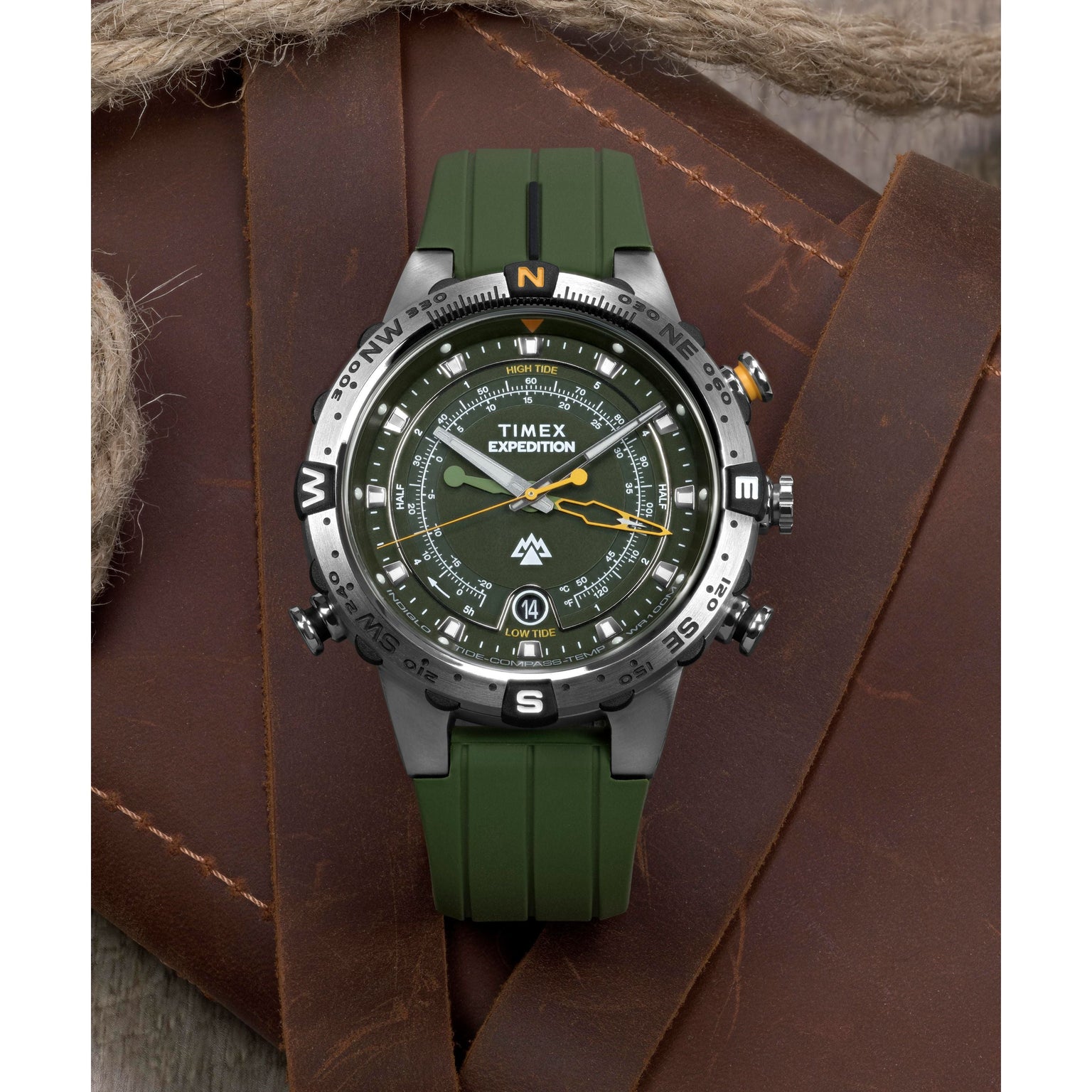 A Timex Expedition® Tide-Temp-Compass analog watch with a green band and black bezel sits on brown leather near thick rope. Its olive-green face features compass markings with white and orange accents.