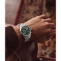 A person wearing a Timex G SS QZ WATRBUY TRAD ACE CH 41 GRN DL 25, featuring a stainless steel case and green face, rests their arm on a brown leather armrest; a patterned rug is partially visible in the background.