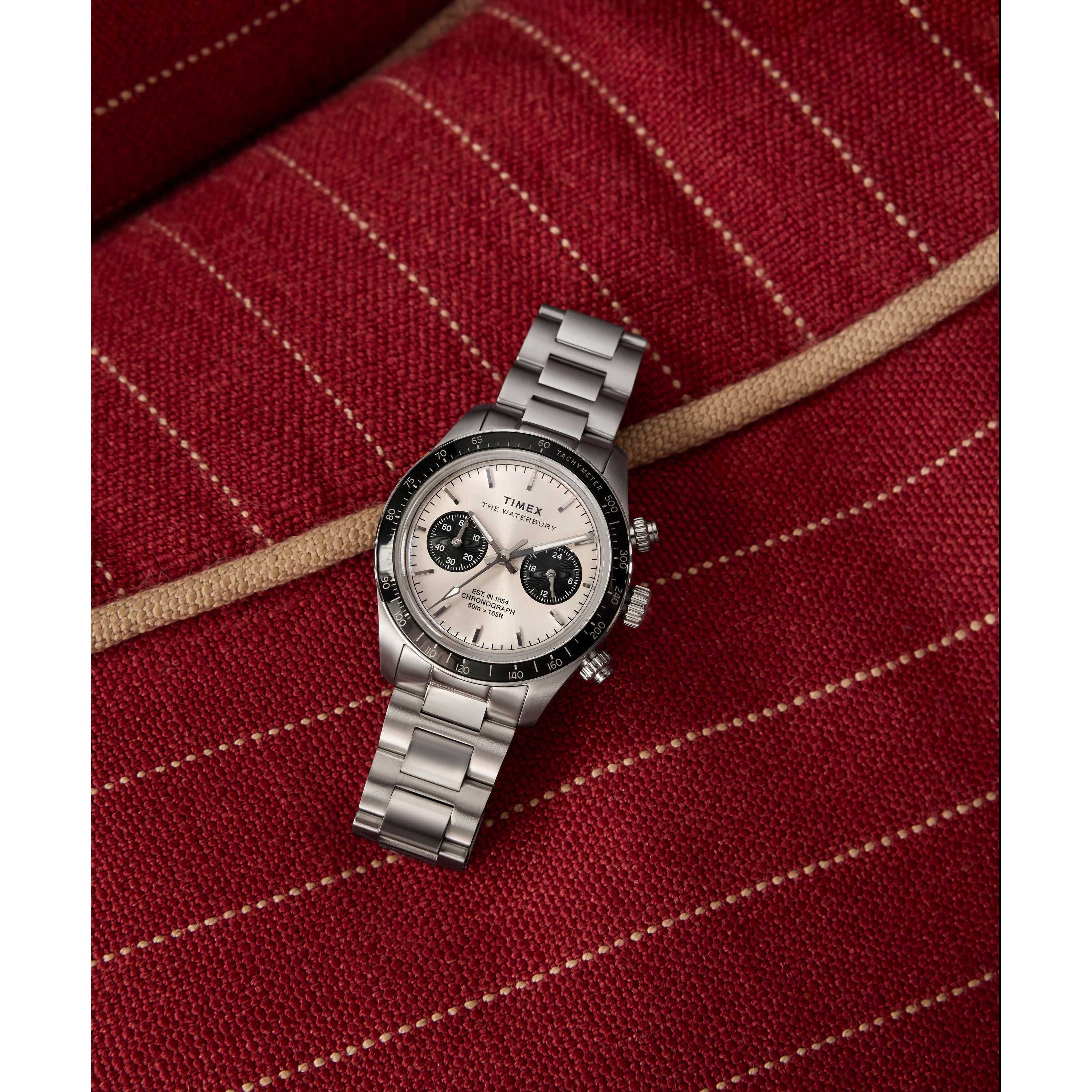 A Timex Waterbury Heritage Chronograph with a stainless-steel bracelet, white dial, and black subdials rests on red, striped, textured fabric.