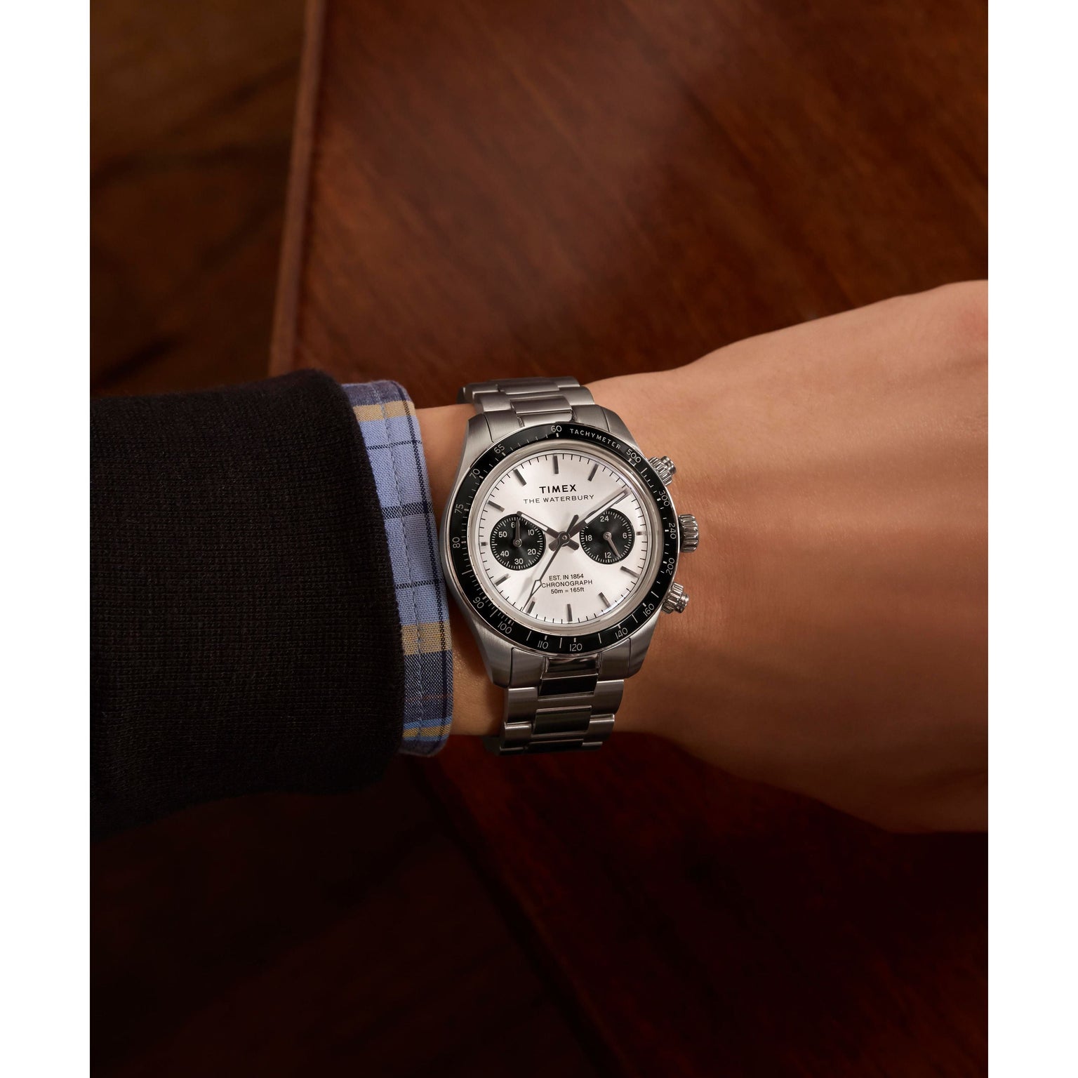 Wearing a Timex Waterbury Heritage Chronograph watch with a stainless-steel bracelet, white face, and black subdials, a person in a black sweater over a blue plaid shirt rests their wrist on a wooden surface.