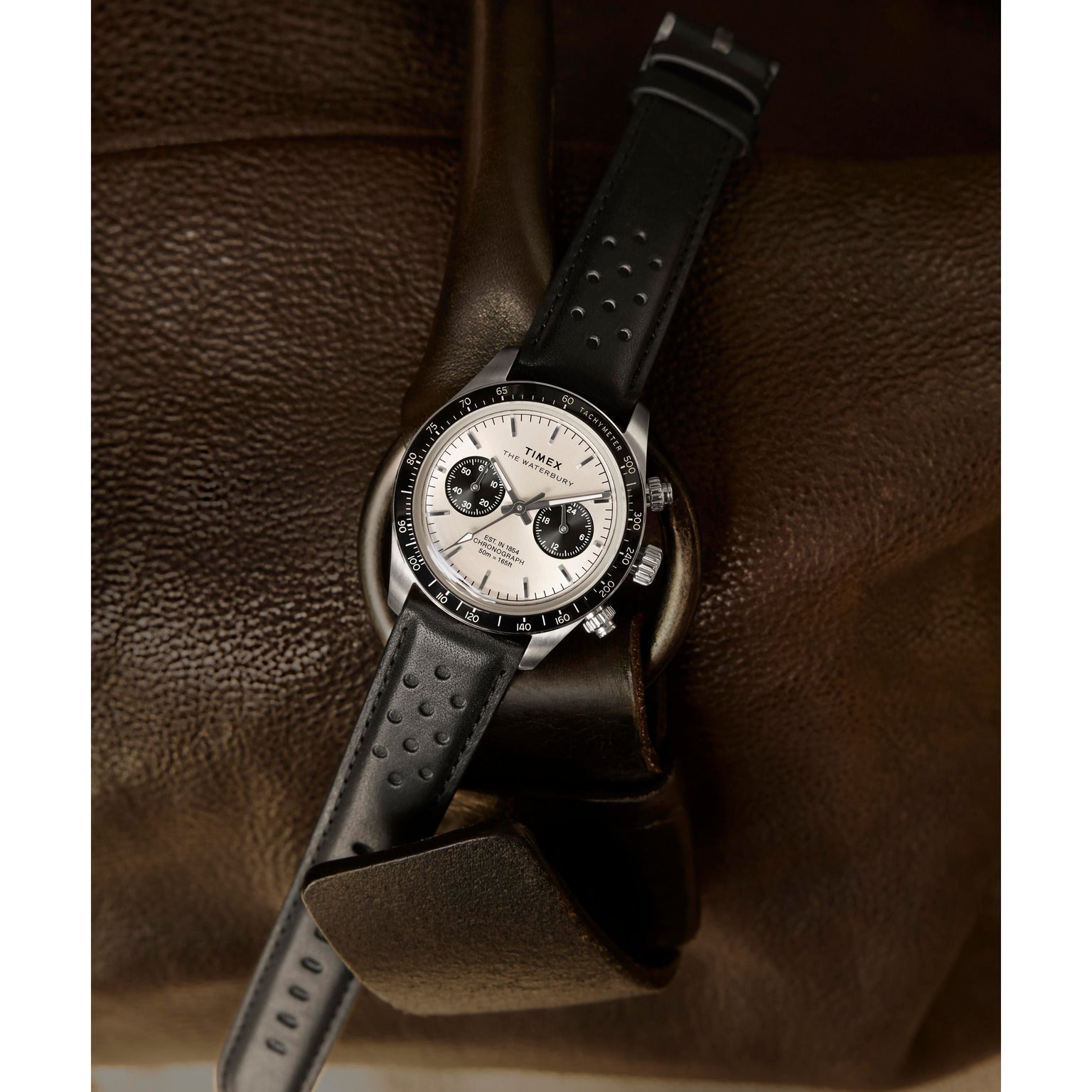 The Timex Waterbury Heritage Chronograph, a silver and black watch with a leather band, rests on brown leather. Its face shows three subdials, a tachymeter bezel, and the time set to about 10:09.