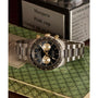 The Timex Waterbury Heritage Chronograph, featuring a silver stainless steel case and black and gold tachymeter bezel with two subdials, is displayed on a green patterned book with blurred books and signs in the background.