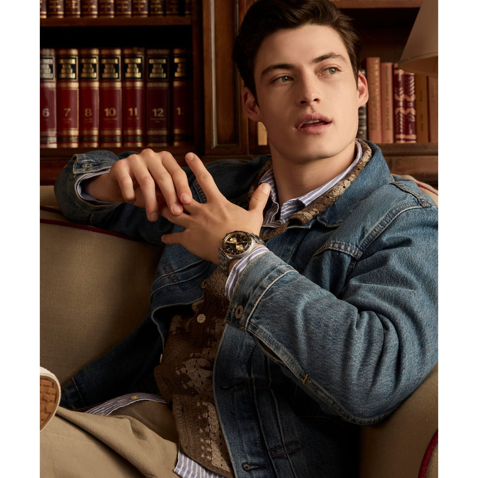 A young man in a denim jacket and a Timex Waterbury Heritage Chronograph sits on a sofa, gazing sideways. Behind him, shelves lined with books evoke a cozy library-like atmosphere.