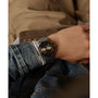 A person wearing the Timex Waterbury Heritage Chronograph, featuring a black dial and tachymeter bezel, styled with a denim jacket and light brown pants, rests their hand on their lap.