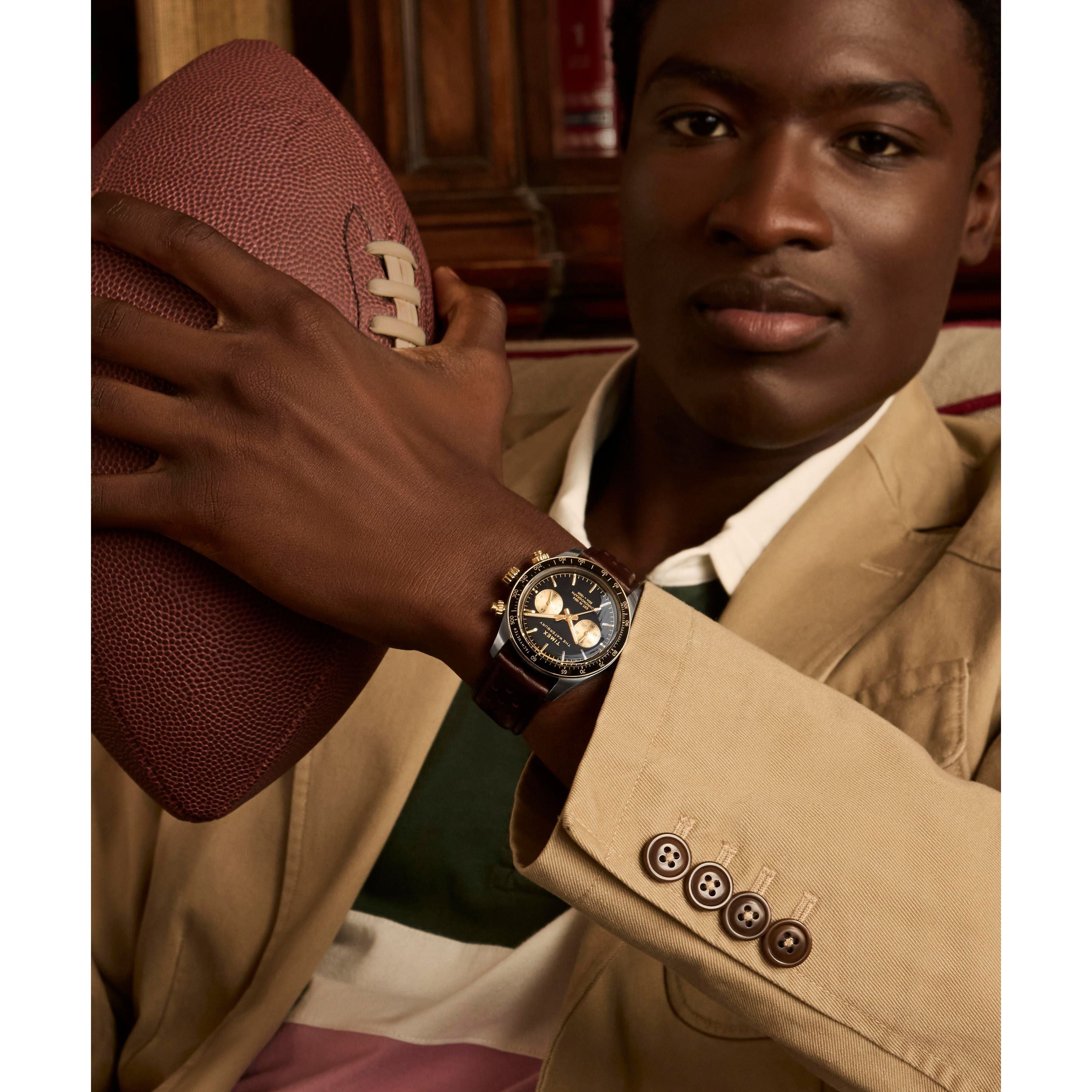 A man in a beige blazer and striped shirt sits holding a football, focused, wearing a Timex Waterbury Heritage Chronograph with a stainless-steel case on his left wrist.