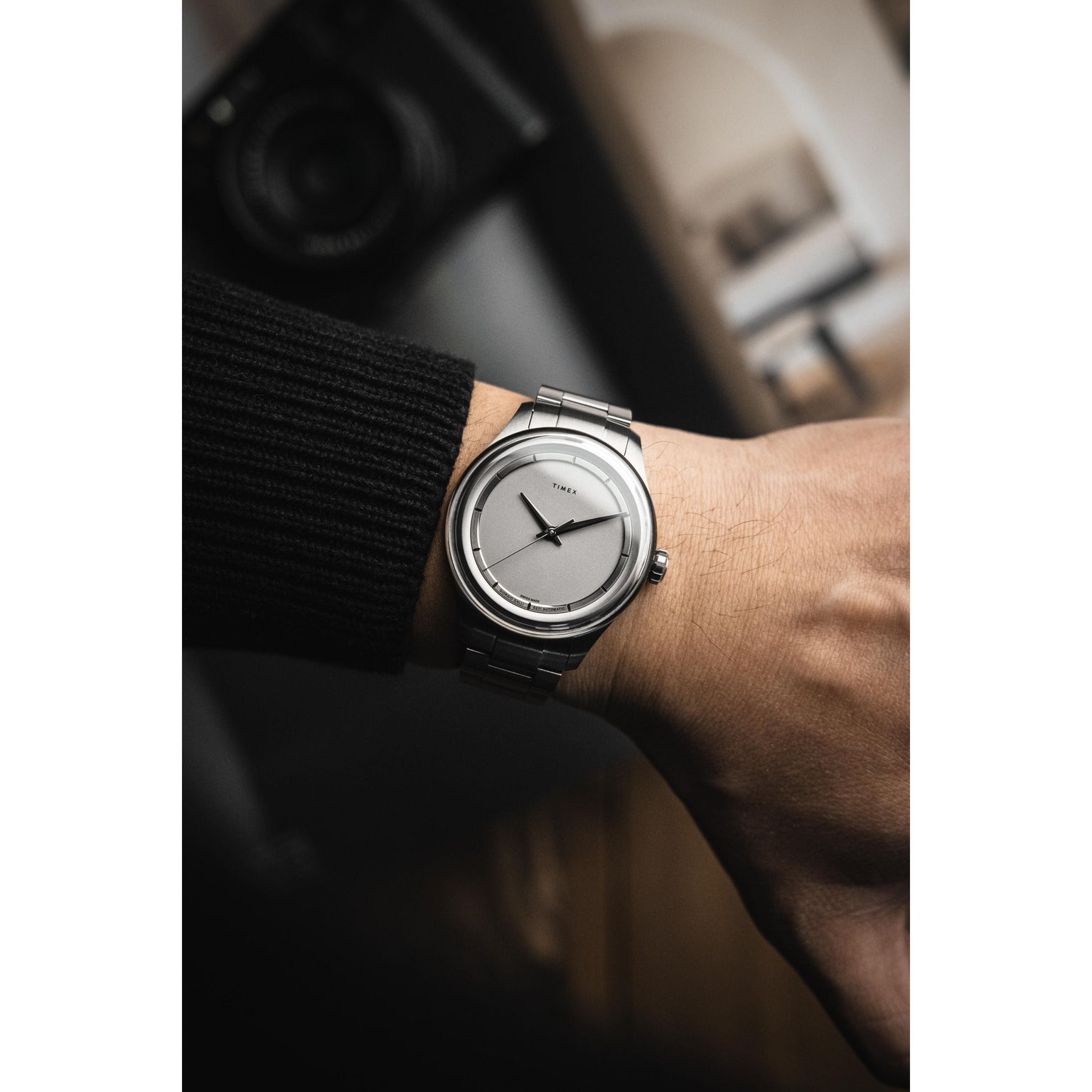 A person wearing a Timex Giorgio Galli S2Ti Automatic, a minimalist 38mm titanium watch with a silver metal band, is viewed from above against a blurred background showing part of a camera and a hand in a dark sleeve.