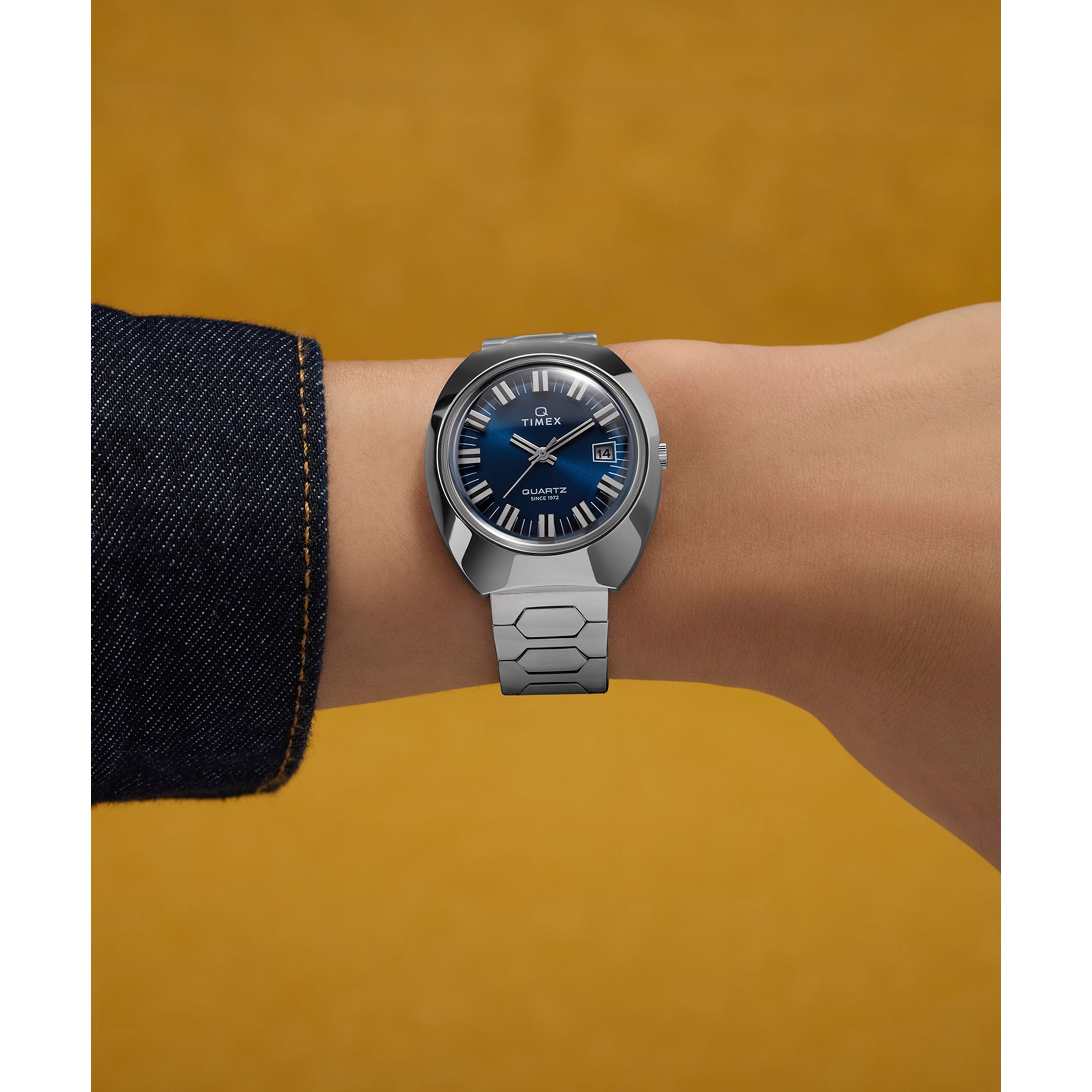 A Timex Q 1972 Time Machine Reissue quartz watch with a blue face is worn on the wrist. The wearer has a dark denim jacket, and the solid mustard yellow background highlights this classic accessory.