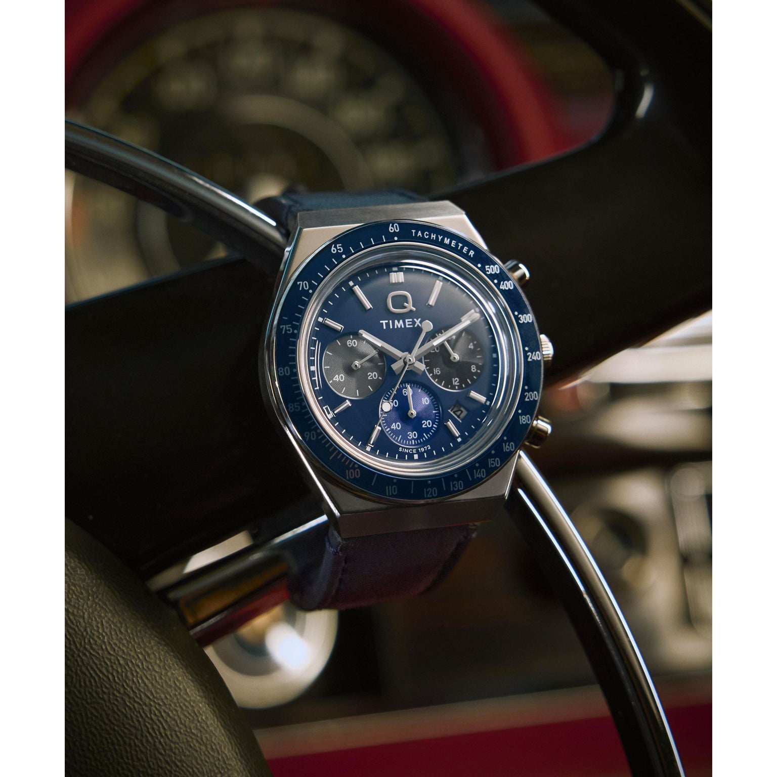 A Timex Q Chronograph with a leather strap and tachymeter bezel is wrapped around a black steering wheel in a classic car, with the blurred dashboard visible in the background.