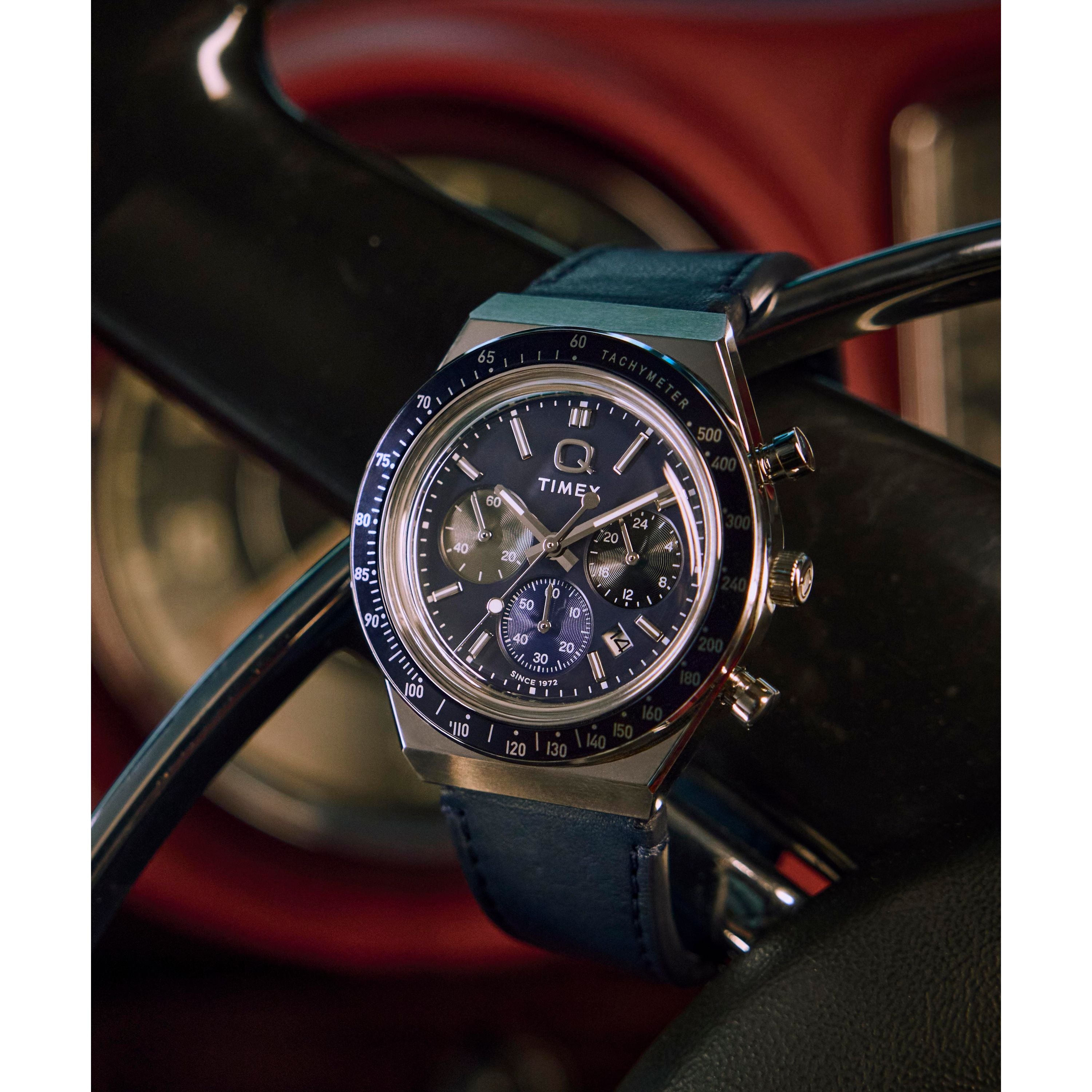A Timex Q Chronograph with a blue strap and multiple dials circles a glossy black steering wheel, its tachymeter bezel gleaming as blurred car gauges disappear in the background.