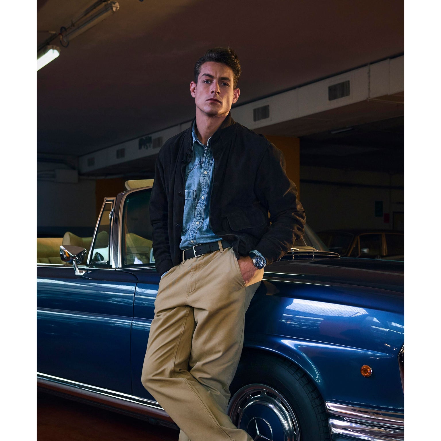 A man in a denim shirt, tan pants, and a dark jacket leans against a classic blue convertible in a dimly lit garage, confidently displaying his Timex Q Chronograph as he looks at the camera with his hands in his pockets.
