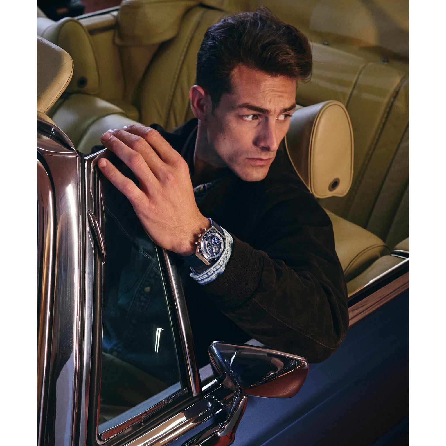 A man with brown hair in a dark jacket, wearing a Timex Q Chronograph watch, sits seriously in the driver’s seat of a classic car with beige leather interior, gazing out the window.