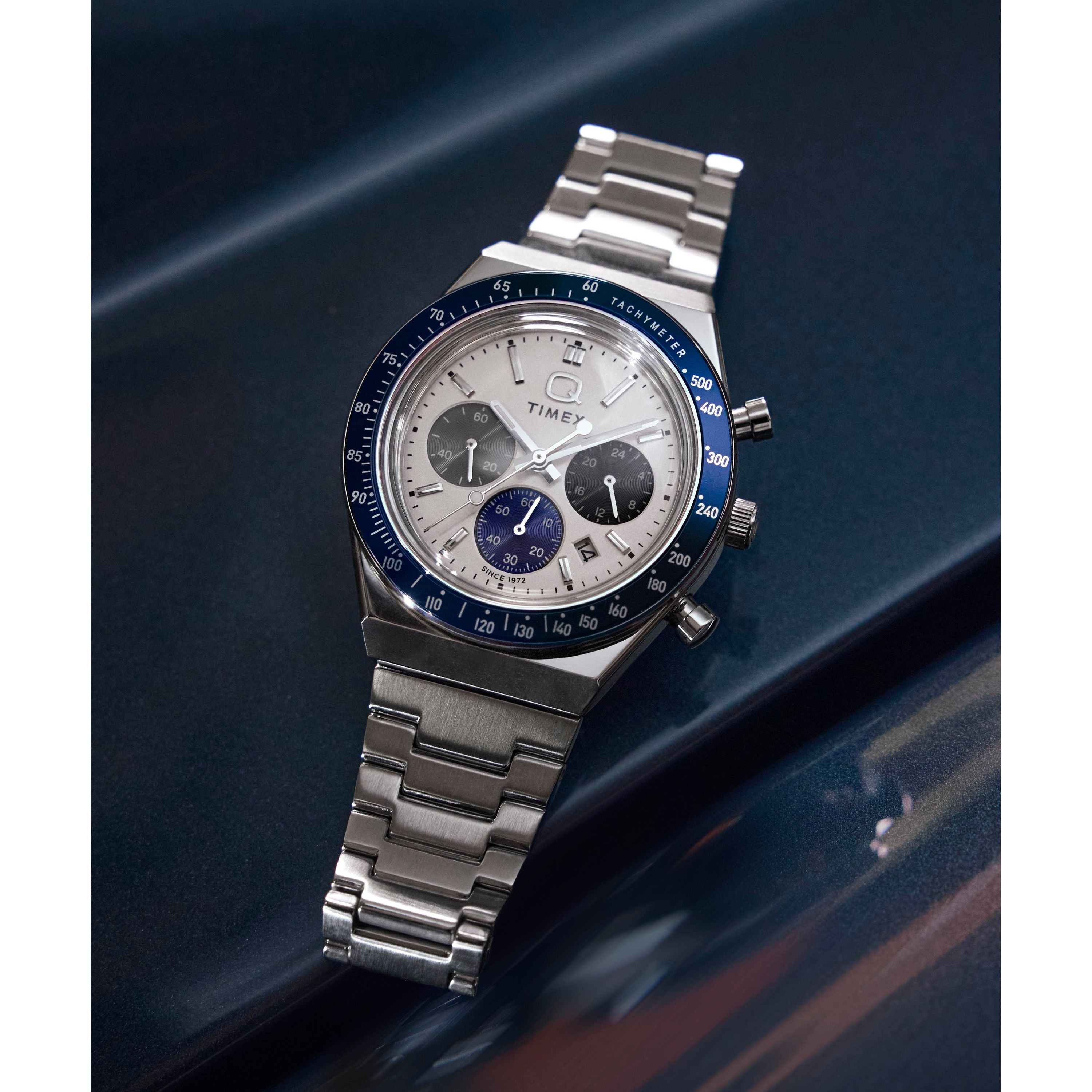 The Timex Q Chronograph is shown on a dark, glossy surface, featuring a stainless steel bracelet, silver dial, blue bezel, three subdials, and three side pushers for precise timekeeping.