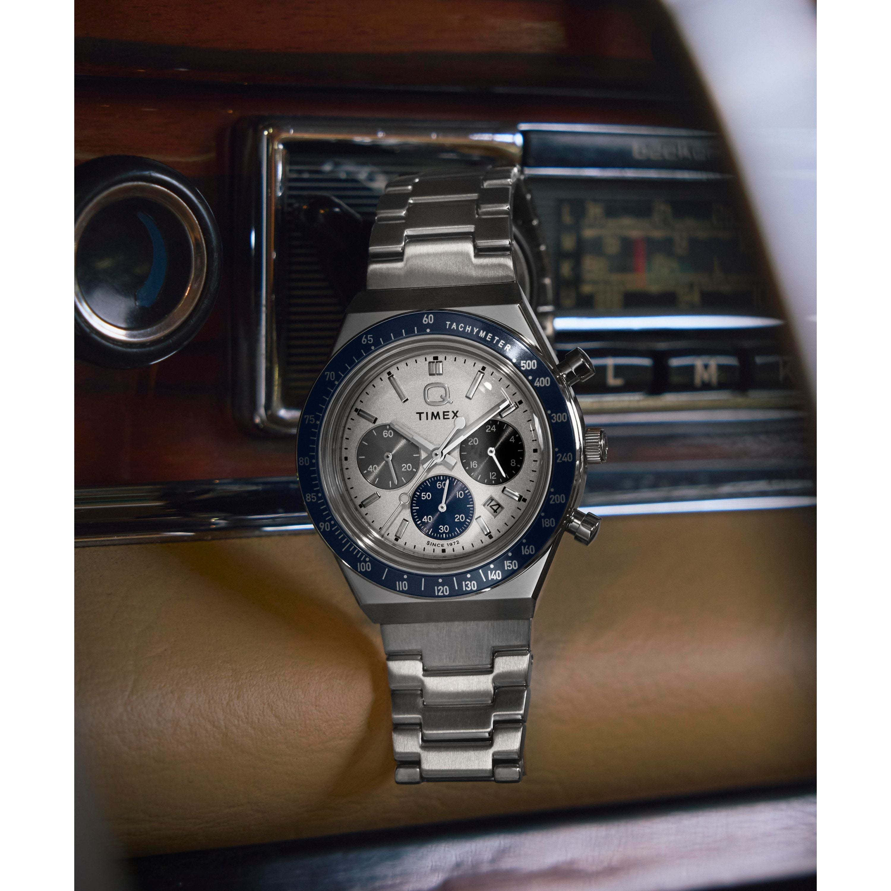 A Timex Q Chronograph watch with a stainless steel bracelet and blue bezel sits on a vintage car dashboard, surrounded by classic dials and a retro radio.