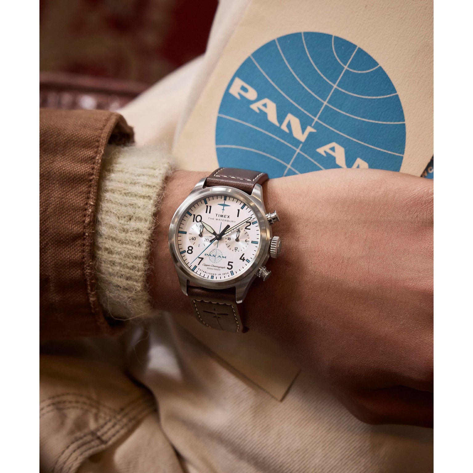 A person wearing the Timex G SS QZ SW WATERBURY TRAD 41 WHITE DL 25, featuring a brown leather strap, stainless-steel case, and white dial, rests their arm on a beige surface while holding a folder with a blue Pan Am logo.