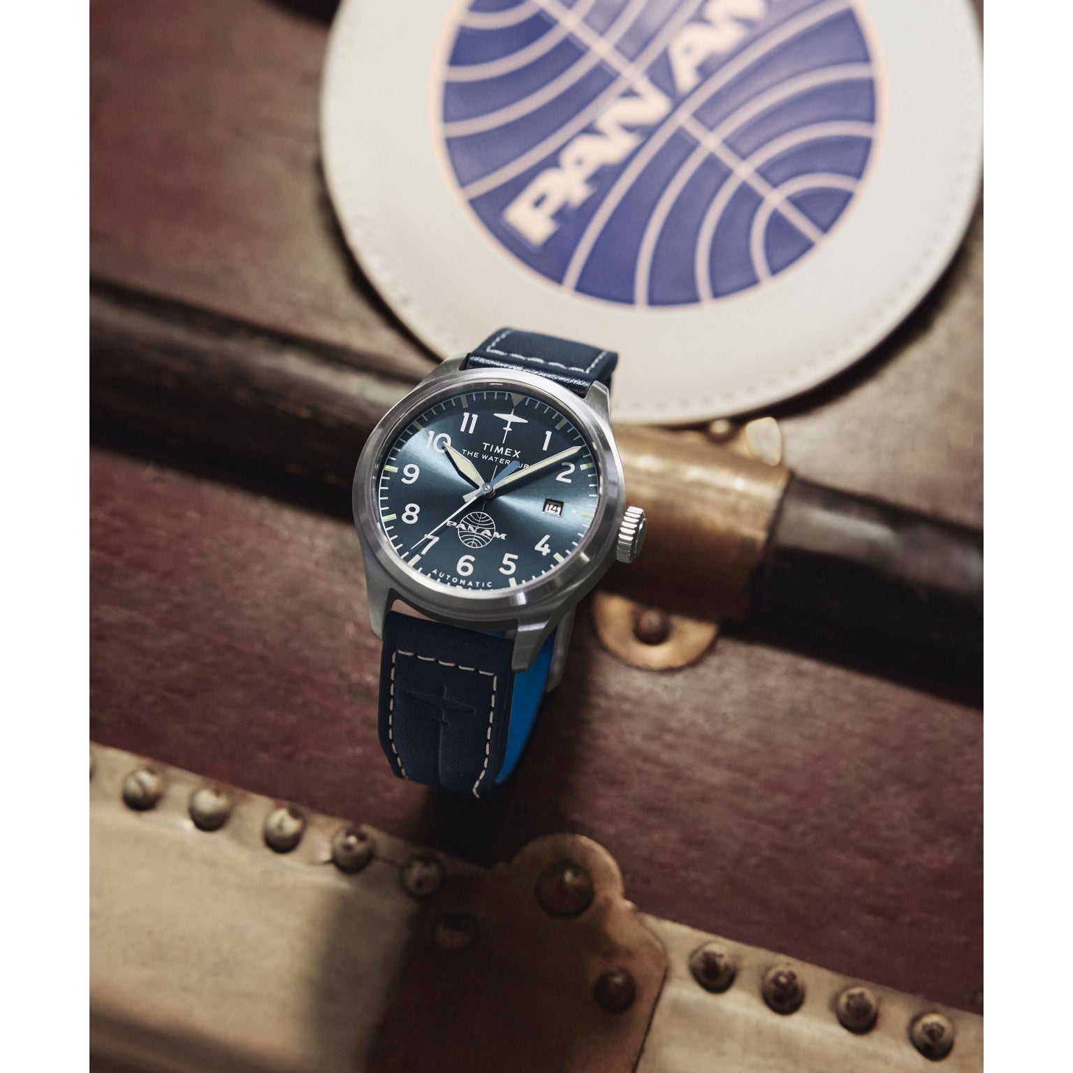 A Timex x Pan Am® Waterbury Automatic Ace watch with a blue strap and stainless steel case sits on a vintage suitcase, with a round Pan Am branded coaster in the background.