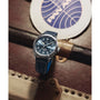 A Timex x Pan Am® Waterbury Automatic Ace watch with a blue strap and stainless steel case sits on a vintage suitcase, with a round Pan Am branded coaster in the background.