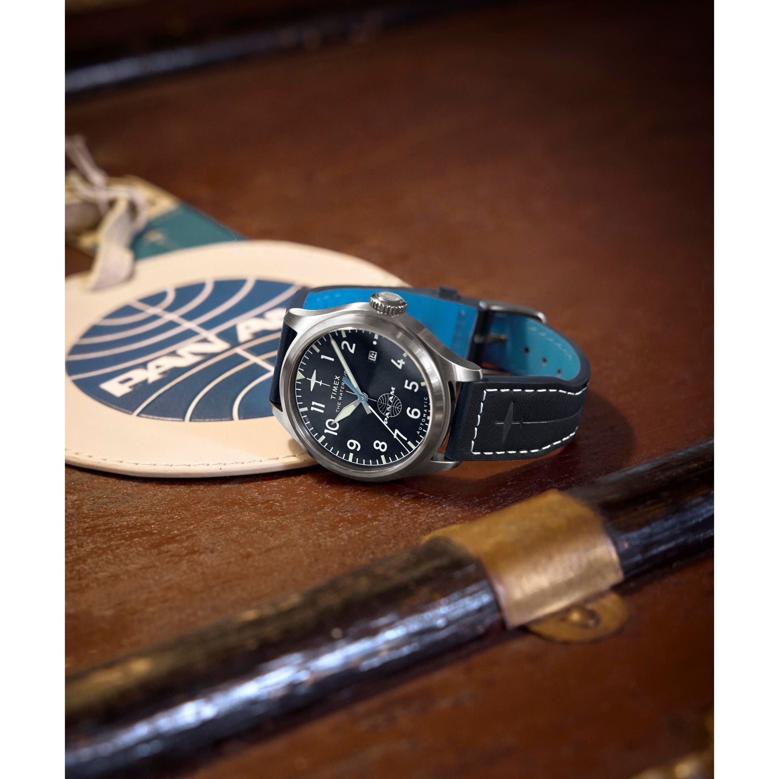 A Timex x Pan Am® Waterbury Automatic Ace watch with a black dial and blue band sits on a vintage suitcase beside a white luggage tag featuring the blue Pan Am logo.