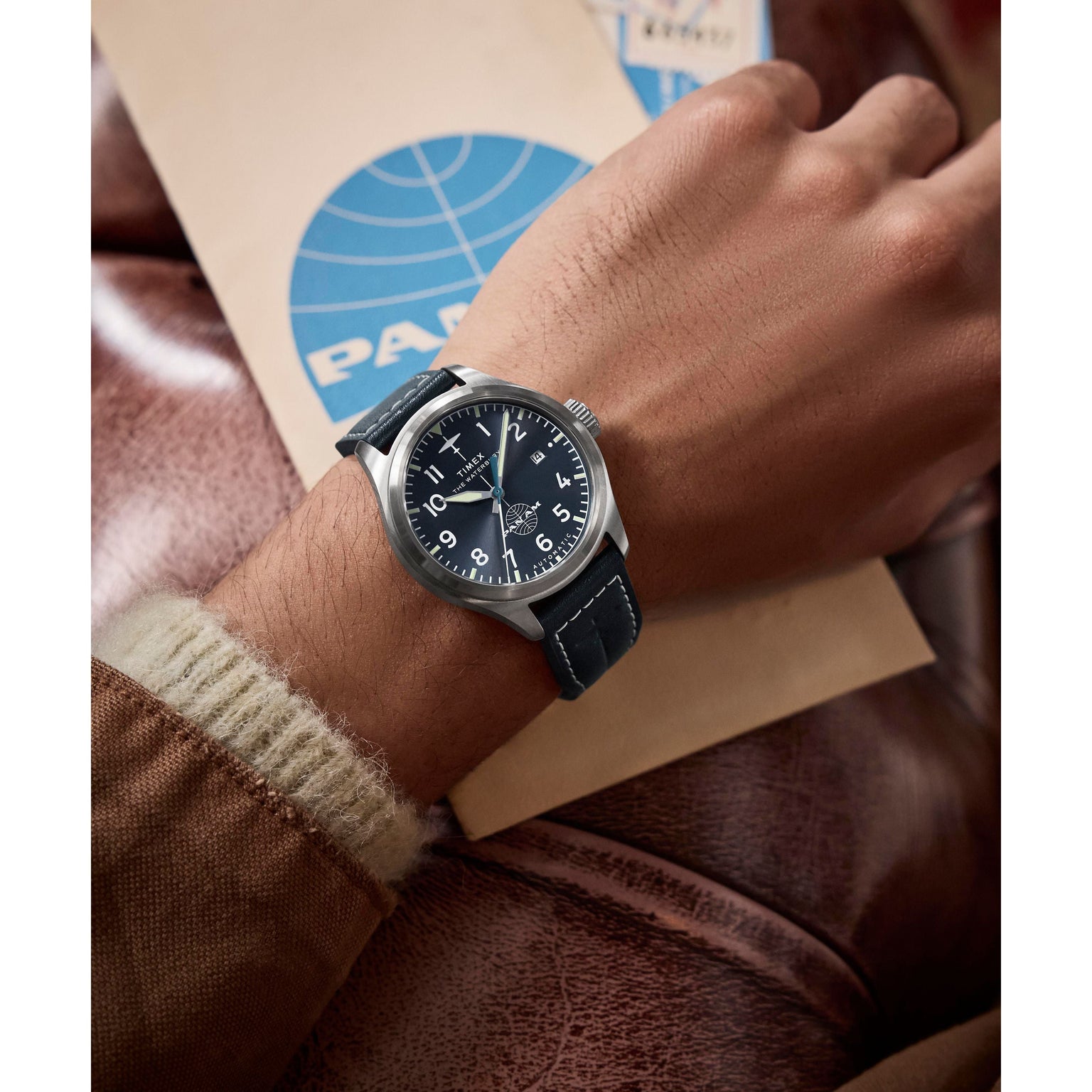 A person wearing a Timex x Pan Am® Waterbury Automatic Ace watch with a black leather strap and stainless steel case rests their hand on a brown leather surface, holding a beige PAN AM envelope with a blue logo.