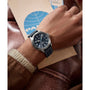 A person wearing a Timex x Pan Am® Waterbury Automatic Ace watch with a black leather strap and stainless steel case rests their hand on a brown leather surface, holding a beige PAN AM envelope with a blue logo.