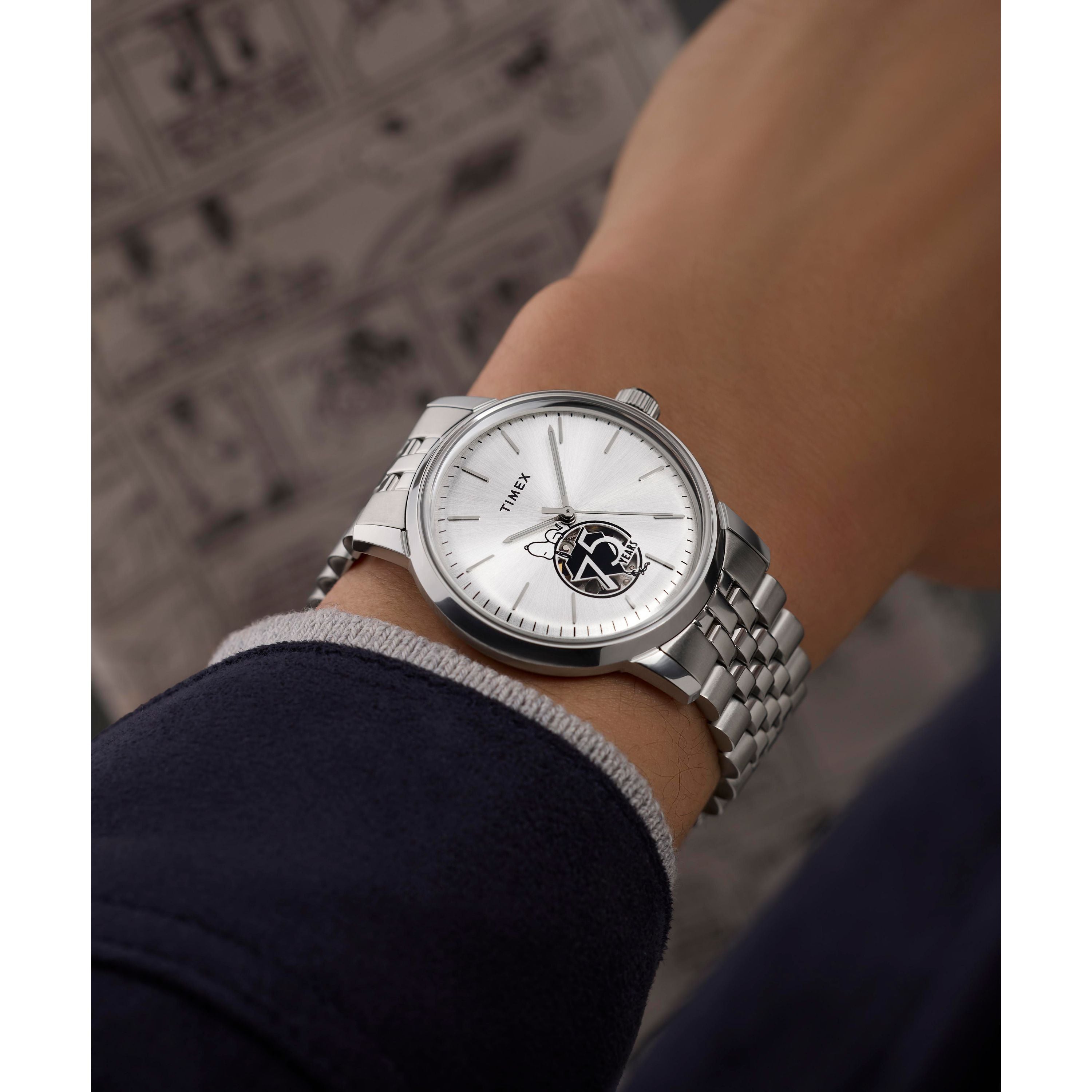 A close-up of a silver Tissot watch with open-heart white dial and metallic band beside the playful Timex G SS AU BW Peanuts Marlin 75 Anni Edi 25, creating an elegant contrast against a dark blue sleeve.