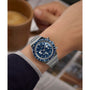 Wearing a Timex x Peanuts® Q Chronograph with a stainless-steel bracelet and blue face, someone holds a cup of coffee as a newspaper sits blurred in the background.