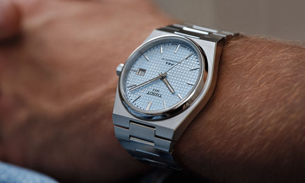 A close up of a person's wrist wearing a Tissot PRX watch with a silver-toned bezel and strap and a light blue textured dial