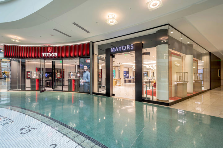 Exterior view of Tudor and Mayors watch stores in a shopping mall, with glass storefronts and displays.