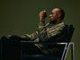 Black sports watch with dark ceramic case worn on wrist of man in olive jacket against green background.