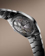 Silver and black automatic watch with exposed mechanism, oval case, and two-tone metal bracelet.