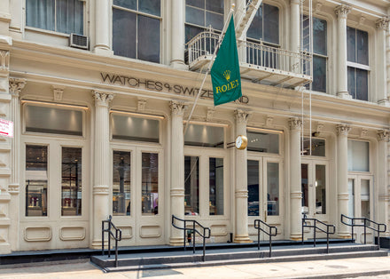 Exterior of a Watches of Switzerland store with a green Rolex flag and steps leading to the entrance.