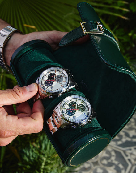 Two luxury wristwatches in a green travel case held outdoors by a person wearing a silver bracelet.