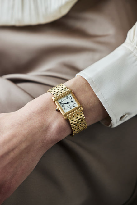A gold wristwatch with a white rectangular face and Roman numerals on a persons wrist.