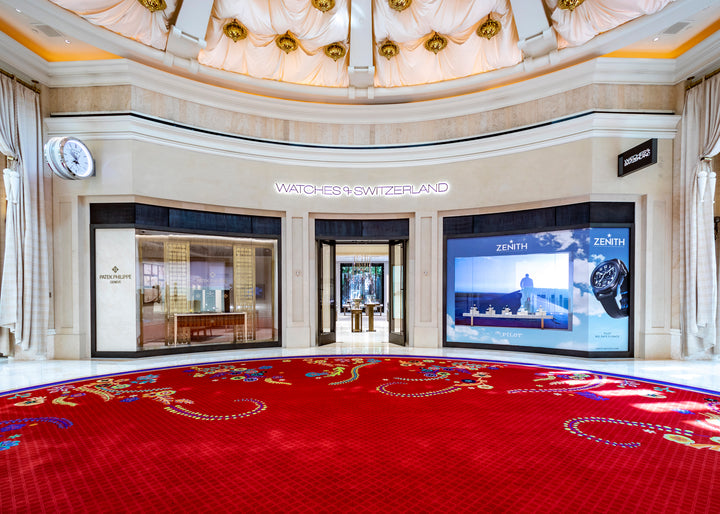 Luxury watch store exterior with bright displays, large screens, and ornate red carpet in front.