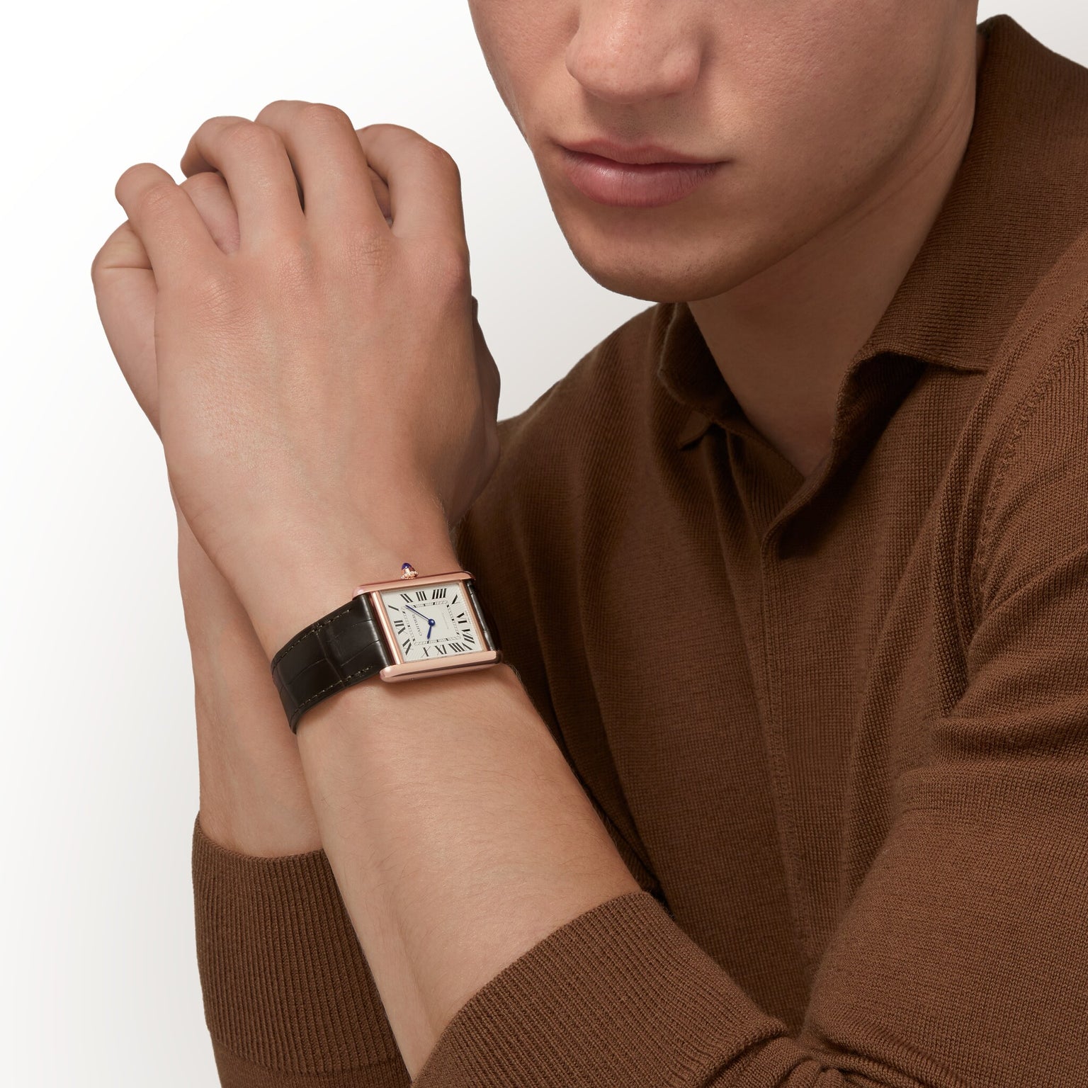 A person in a brown long-sleeve shirt rests their hands together, showcasing the Cartier Tank Louis Cartier Large Model with a white dial, black Roman numerals, and a black leather strap—a true tribute to timeless design.