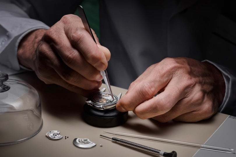 A watchmaker uses tweezers to repair a watch mechanism on a workbench with tools nearby.