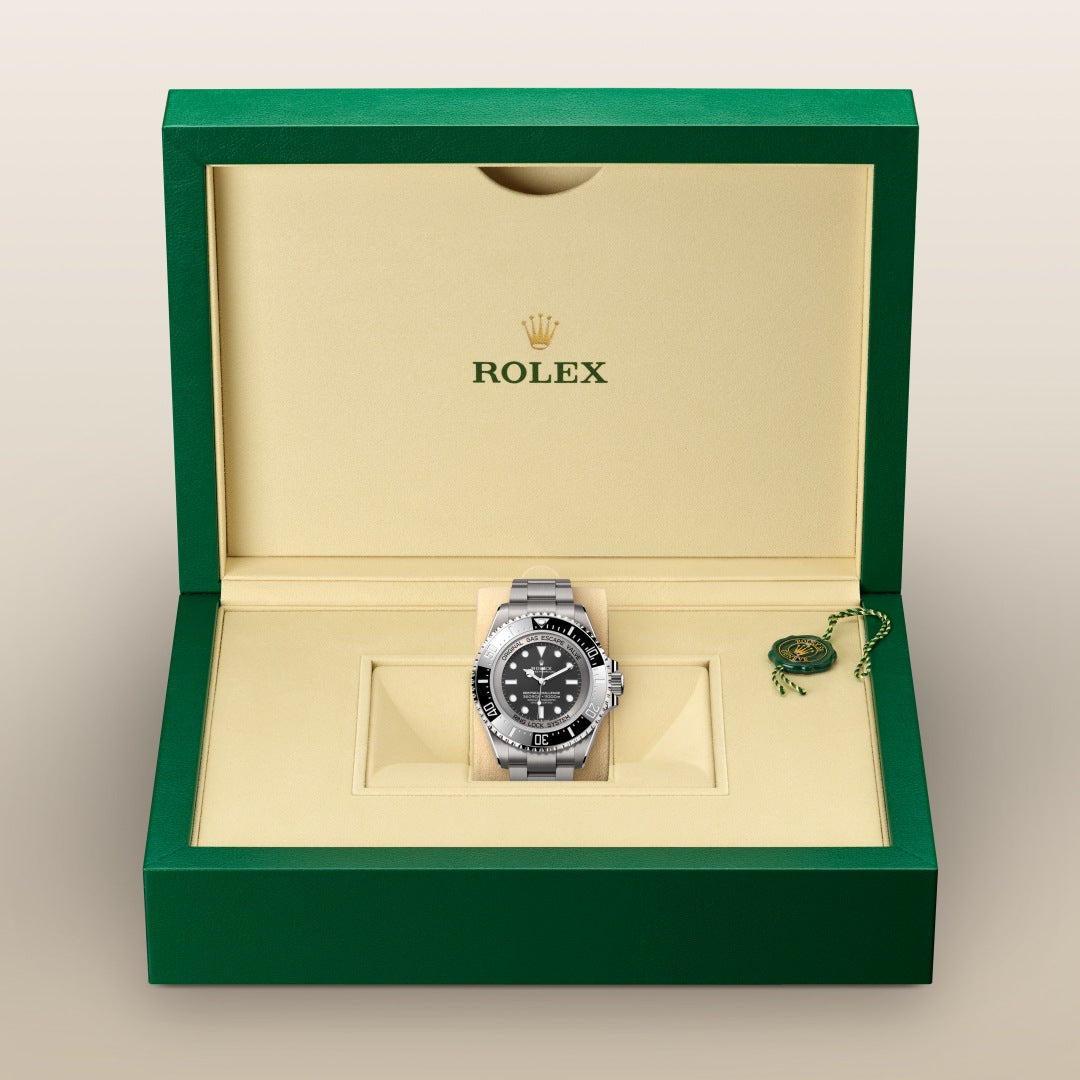 A Rolex watch in a green and cream box, with a black dial and silver bracelet, and a green tag beside it.