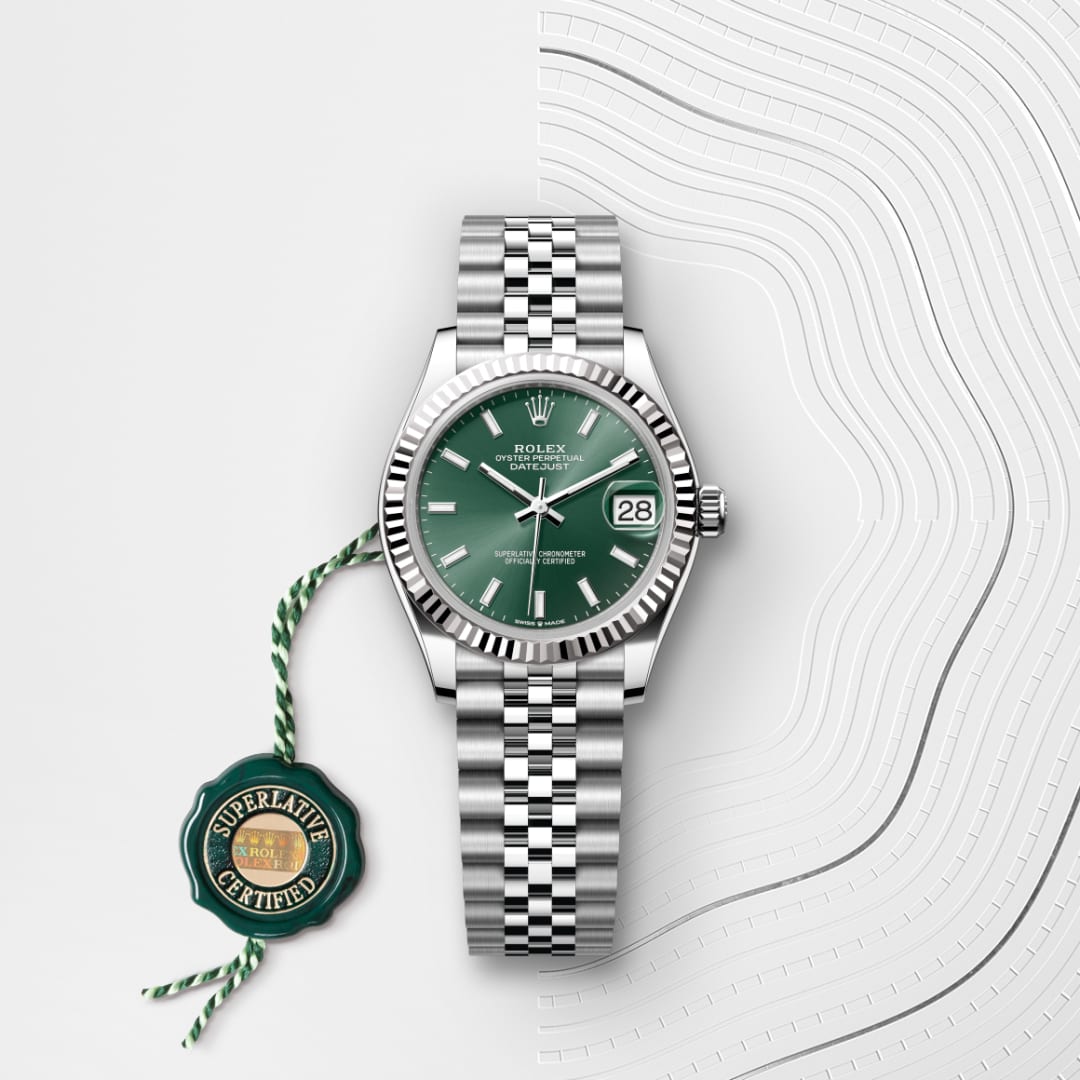 A Rolex watch with a green dial and silver bracelet, next to a green Superlative Certified tag on a textured surface.
