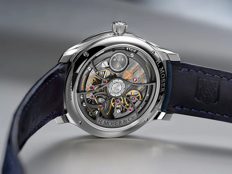 Stainless steel watch with exposed skeleton dial showing gold and silver mechanical movement, dark blue leather strap.