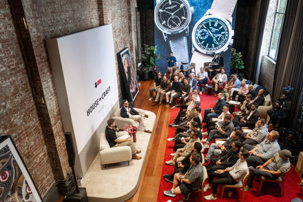 Two people speak onstage at a watch event as an audience listens; large watch images are displayed behind them.